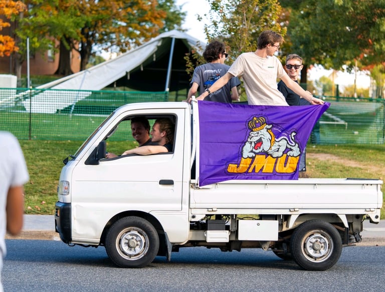 Luke Gilmore riding on the back of a Kei truck with a JMU banner on it.