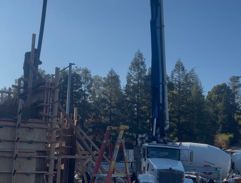 A white concrete boom pump truck and mixer pouring cement into wooden wall forms on a construction site.