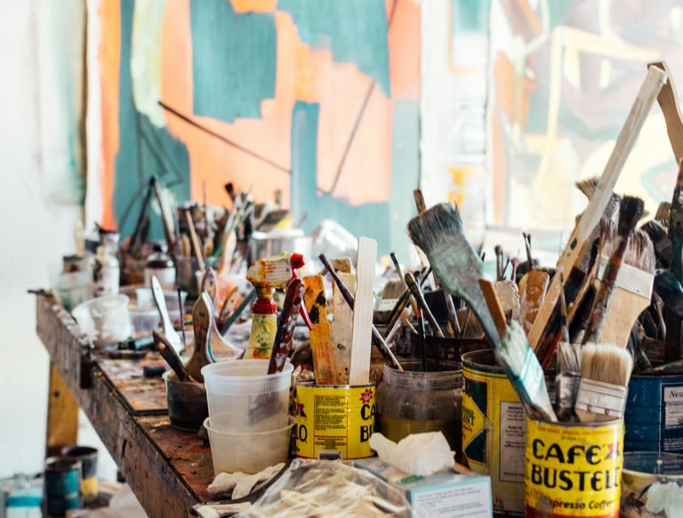 A collection of different sized used artist paintbrushes sit on a paint stained table.