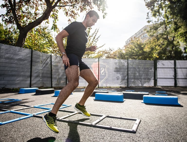 A man performs agility ladder drills and box jumps during an outdoor fitness workout in a sunny park.