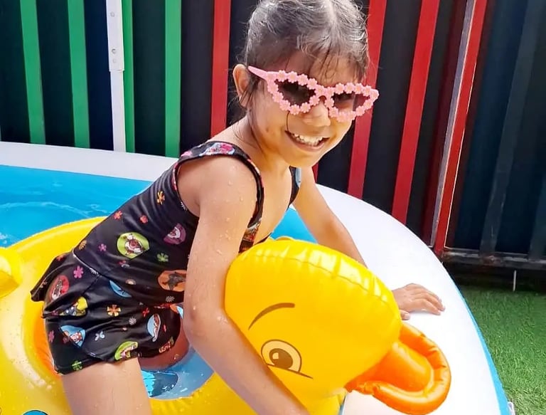 A happy toddler wearing pink sunglasses and a swim vest playing in a pool at Enfants Preschool 