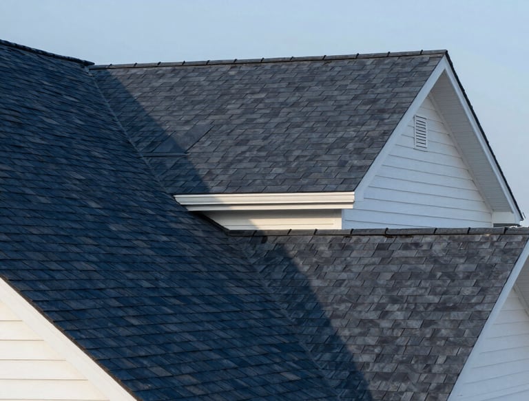 A wide-angle, professional photograph of a beautifully shingled residential roof in a North American / US suburban neighborhood. The shingles are a crisp Dark Navy, contrasting with the Off-White exterior of the house. The sky is a clear Soft Blue, suggesting a reliable and sturdy structure under bright daylight.