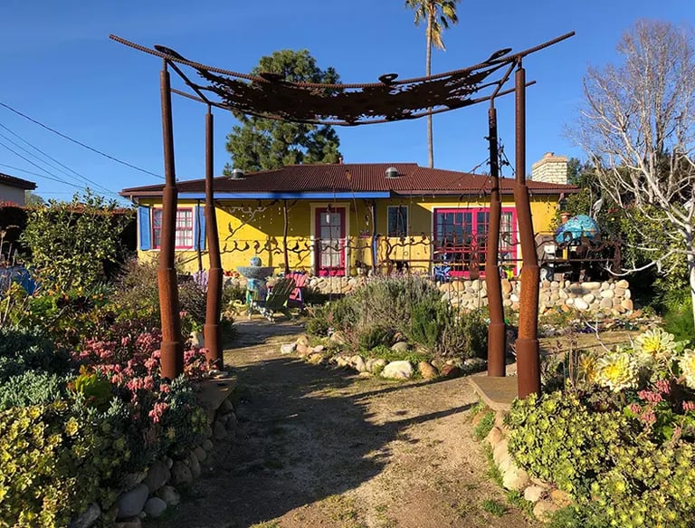 Yellow Santa Barbara home with pink trim and blue shutters, viewed through a rustic metal garden arbor.