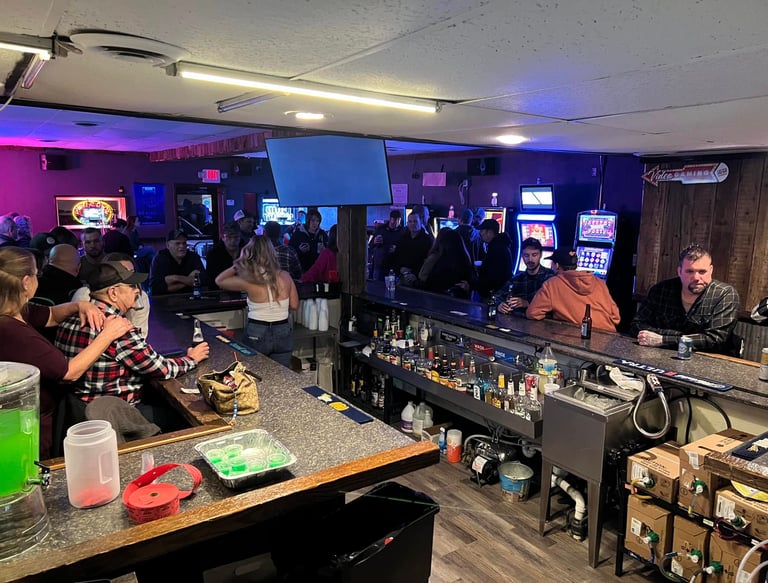 Crowded local bar with patrons, slot machines, and bartenders serving drinks in a dimly lit tavern.