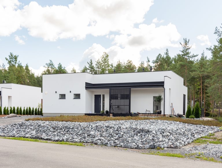 Modern white minimalist villa with a flat roof and stone landscaping surrounded by a pine forest.