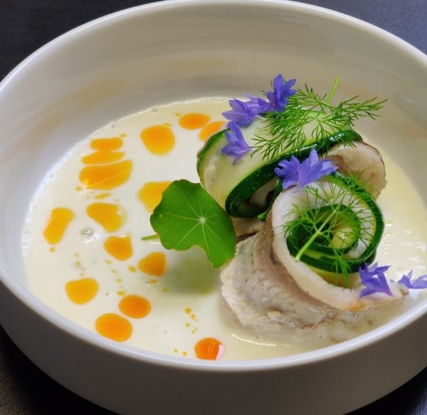 Elegantly plated fish dish with courgette, blue edible flowers, dill, and saffron oil in a white bowl