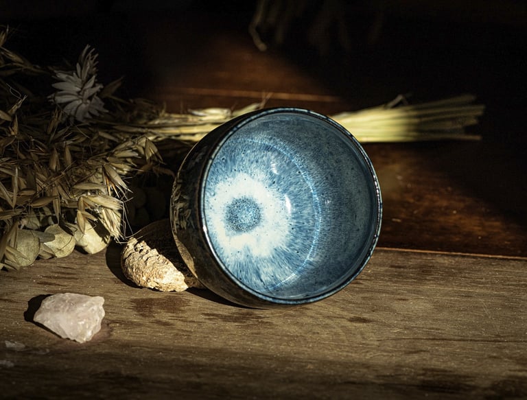 A handmade blue glazed ceramic tea bowl on a rustic wooden table with dried botanicals.
