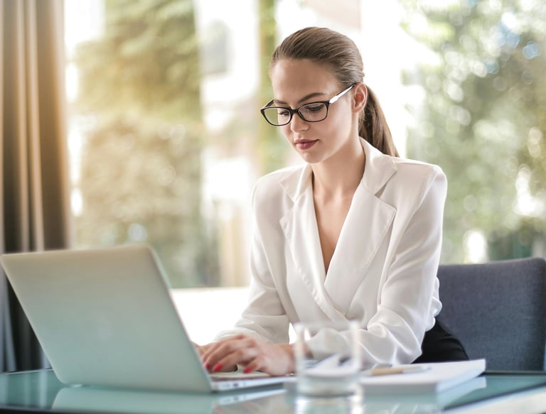 a woman in a white shirt and glasses is sitting at a table and working on laptop