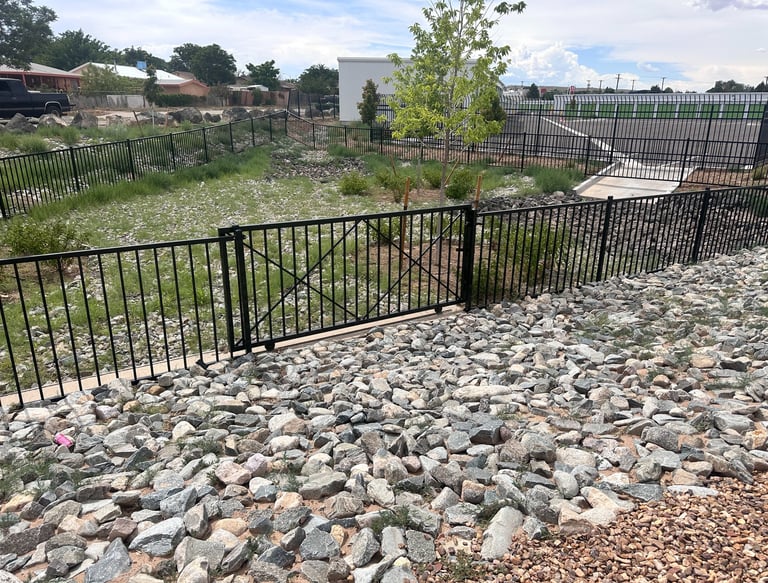 Black decorative metal security fence installed around a landscaped drainage basin with grey river rock.