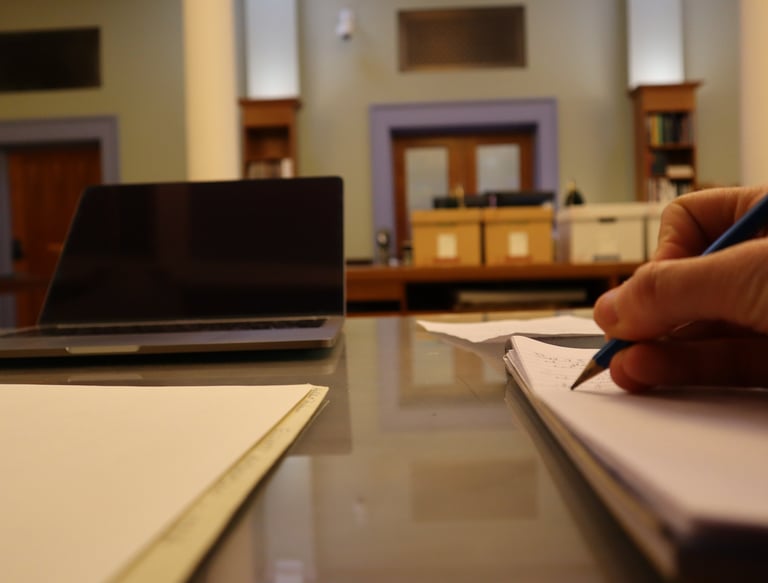 View of a desk in an archival research room showing a person writing in a notebook.