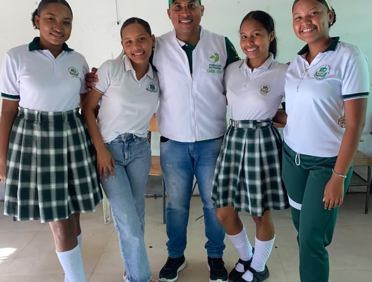Smiling students in green plaid school uniforms pose with a male mentor in a white classroom.