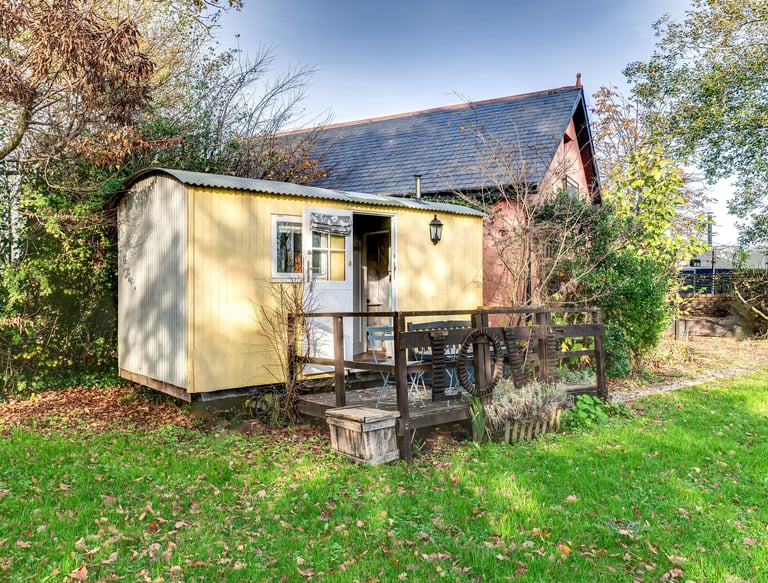 A rustic yellow shepherd's hut with a wooden deck and table set in a green garden.