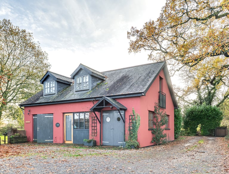 A red cottage with grey shutters and dormer windows nestled among autumn trees and fallen leaves.