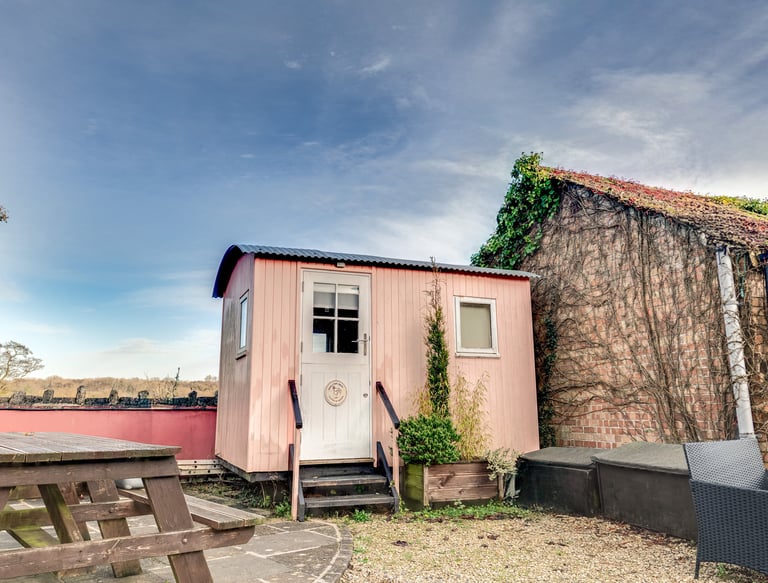 A pink shepherd's hut with a wooden picnic table and rustic brick wall.