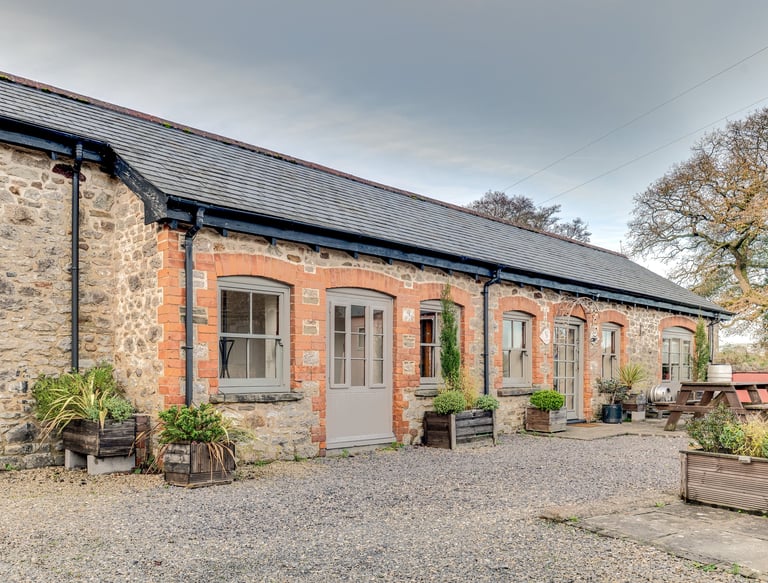 A rustic converted stone barn featuring grey wooden doors and window frames with a gravel patio.
