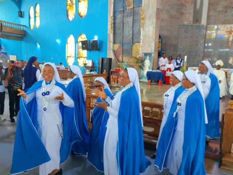 Smiling Catholic nuns in blue and white habits singing during a church service at a cathedral.