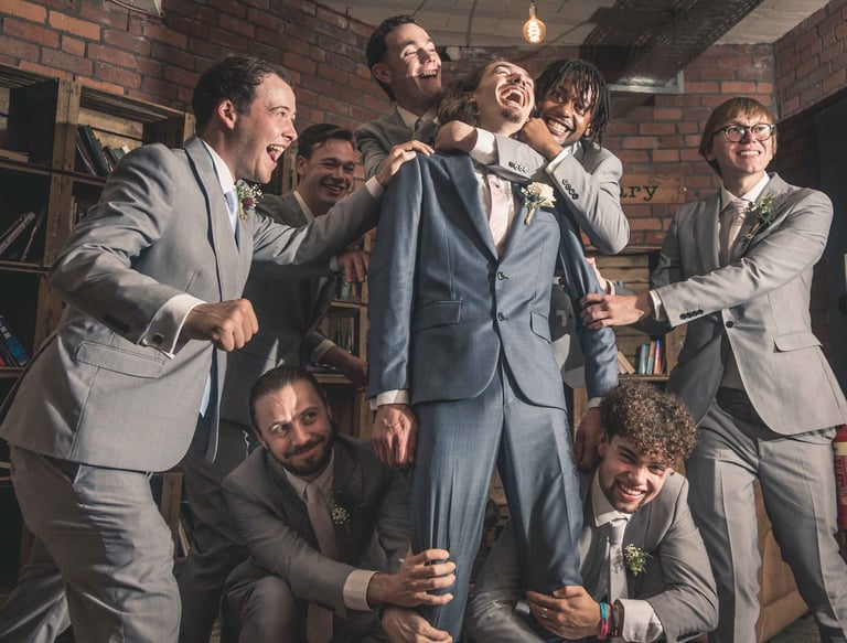 Groomsmen in grey suits laughing and lifting the groom in a blue suit during wedding celebrations.