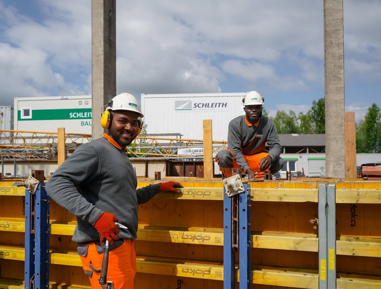 Zwei Bauarbeiter in Schutzkleidung und Helmen arbeiten lächelnd auf einer Baustelle.