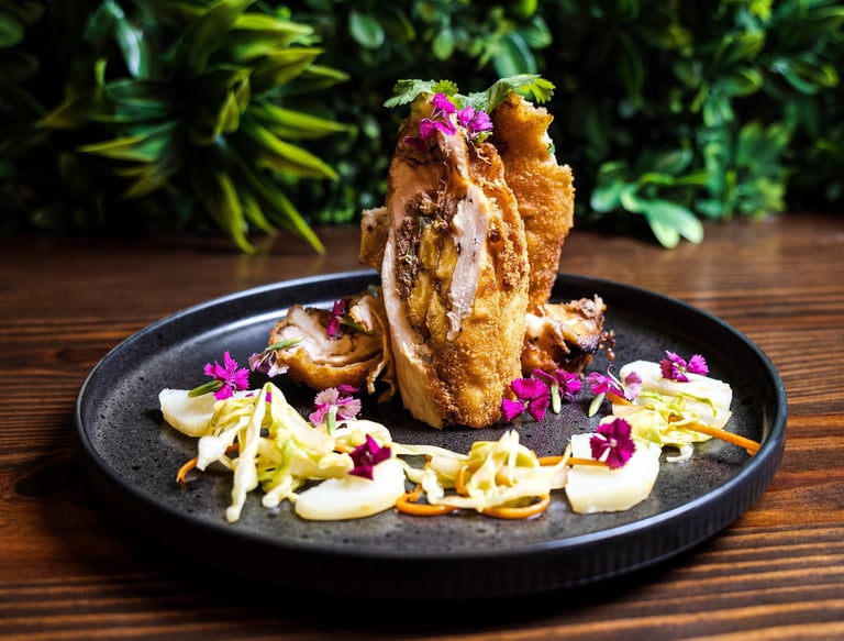 Gourmet stuffed chicken rollatini with plantains, garnished with edible purple flowers and slaw on a black plate.
