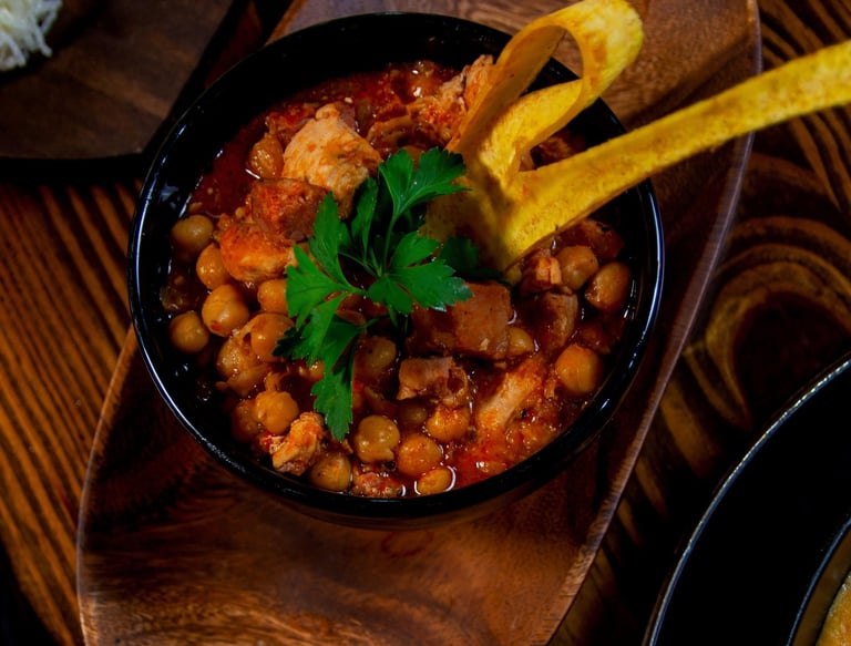 A bowl of Spanish chickpea stew with pork and plantain chips on a rustic wooden tray.