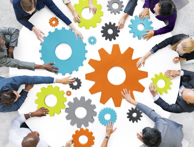 A group of people seated around a table are collaboratively arranging colorful gear-shaped pieces, symbolizing teamwork