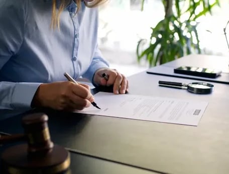 Manos firmando un documento en el escritorio con lupa y carpeta.”