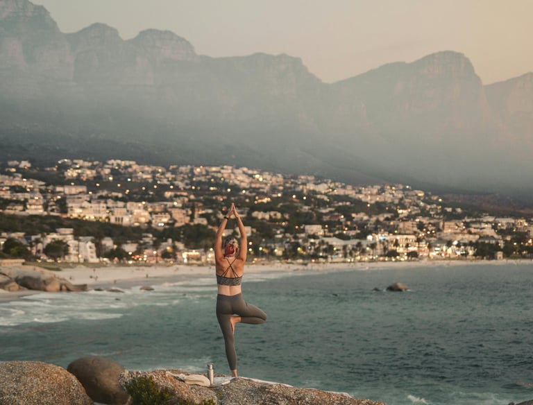 Woman doing yoga on a rocky beach with a coastal city and mountains in the background.
