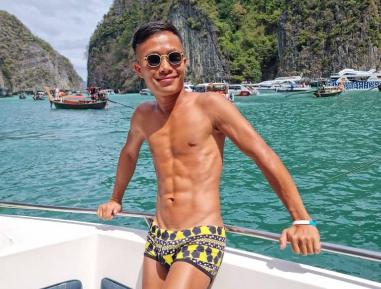 A male outcall masseur in pattaya standing on a boat wearing swim wear with the sea and an island in the background