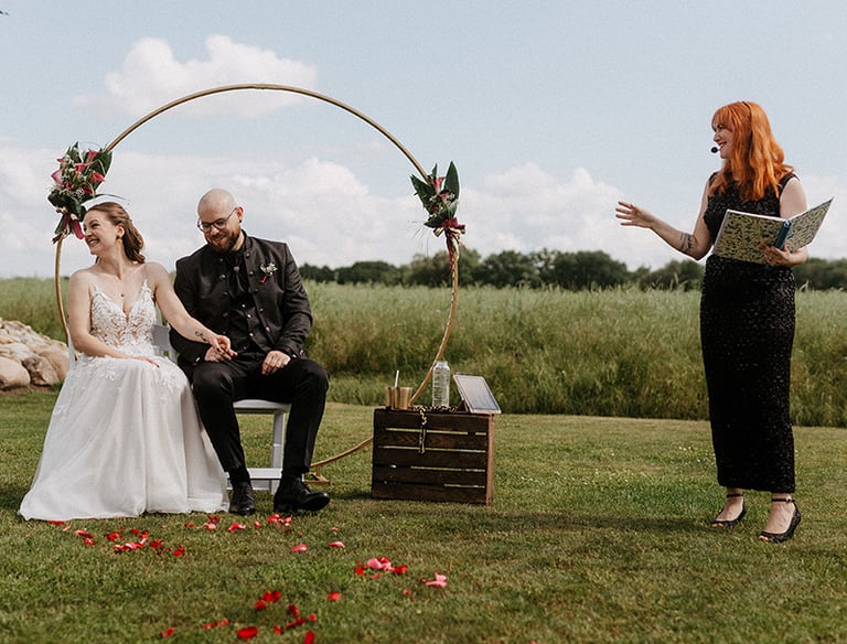 grinsend sitzen Braut und Bräutigam nebeneinander während die Traurednerin im schwarzen Glitzerkleid eine Anekdote erzählt
