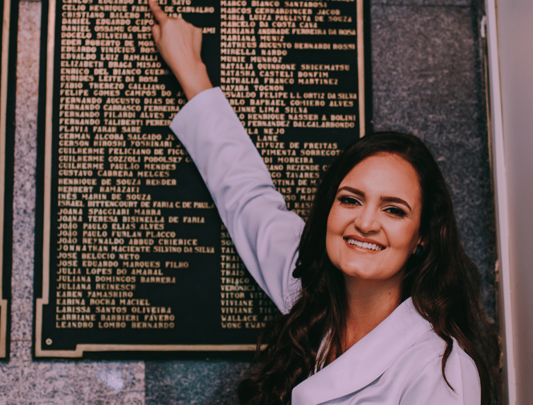 Dra Bruna Melchior de jaleco branco apontando seu nome gravado em placa metálica no hospital das clinicas da USP de ribeirao