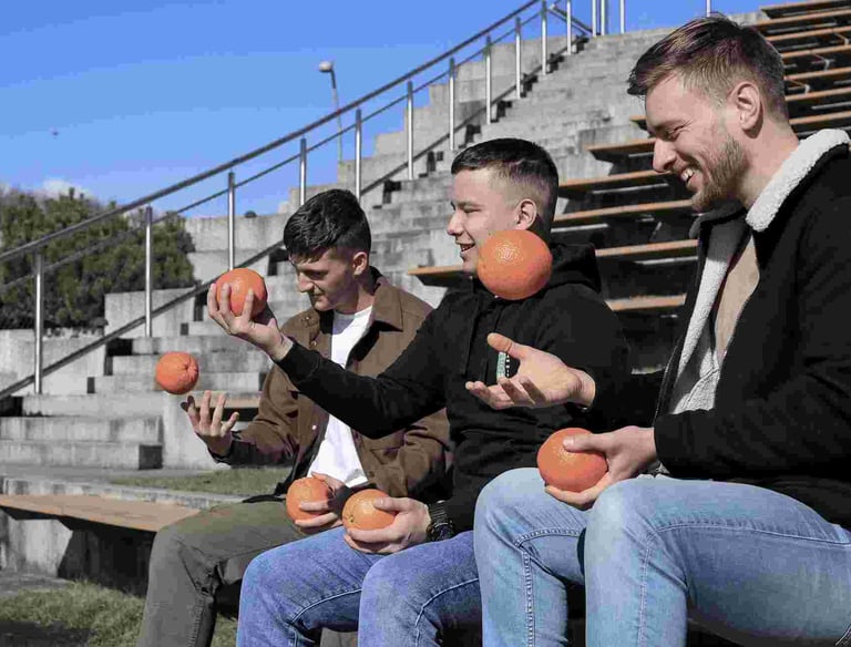 Trys jauni vyrai sedi ir meto apelsinus