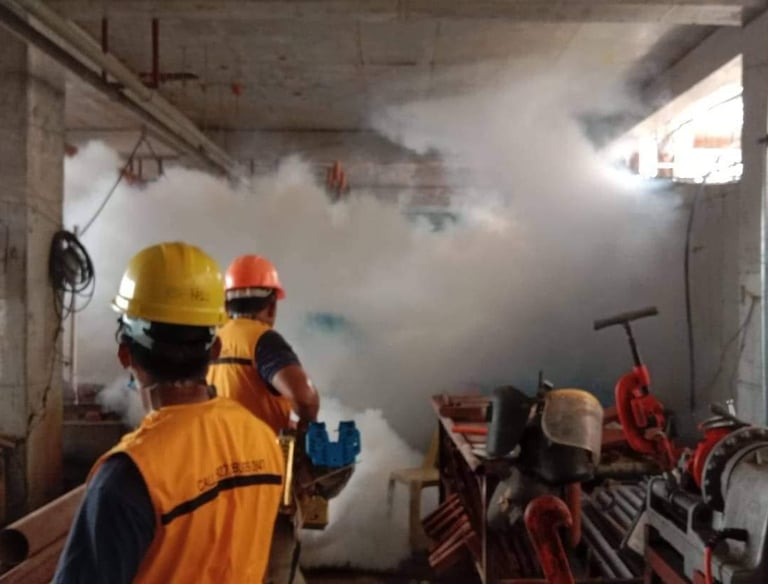 Construction workers in safety vests and hard hats monitoring a fogging machine inside a building site.