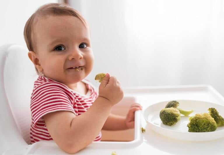 Un bebé sonriente en una silla alta comiendo brócoli fresco al vapor durante la alimentación