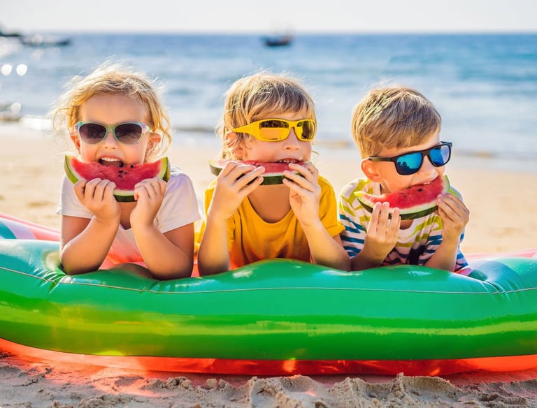 Three young children wearing sunglasses eating fresh watermelon slices on a sunny beach.