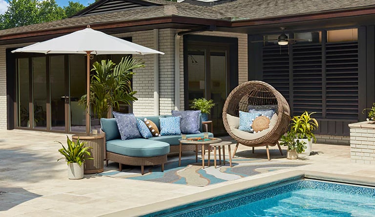 Modern poolside patio featuring a blue outdoor sectional sofa, wicker pod chair, and white sun umbrella.