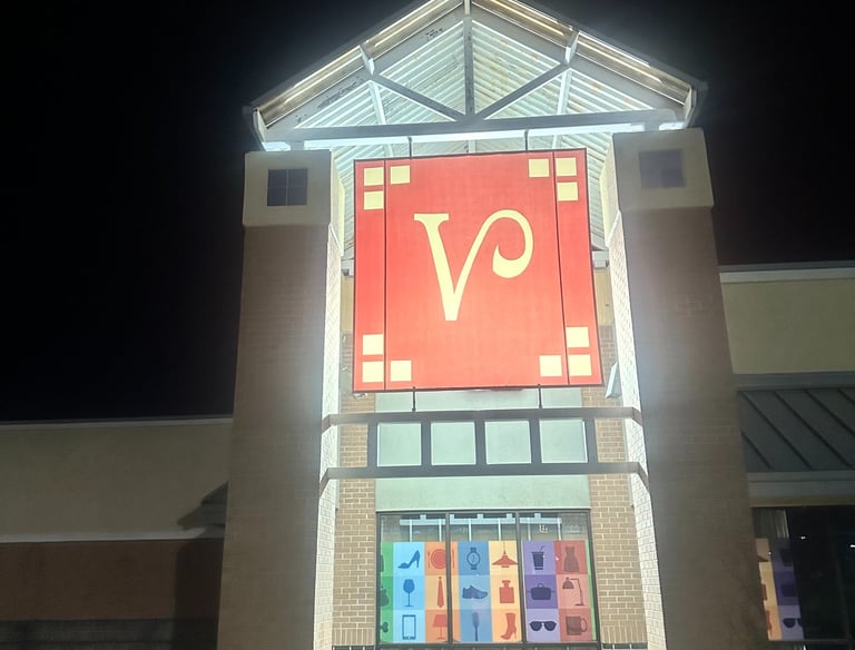 Illuminated shopping mall entrance at night featuring a large red logo sign and retail category icons.