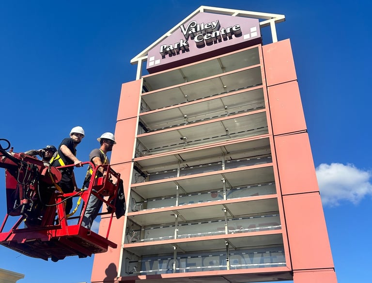 Technicians in a bucket lift repairing the Valley Park Centre pylon sign under a clear blue sky.