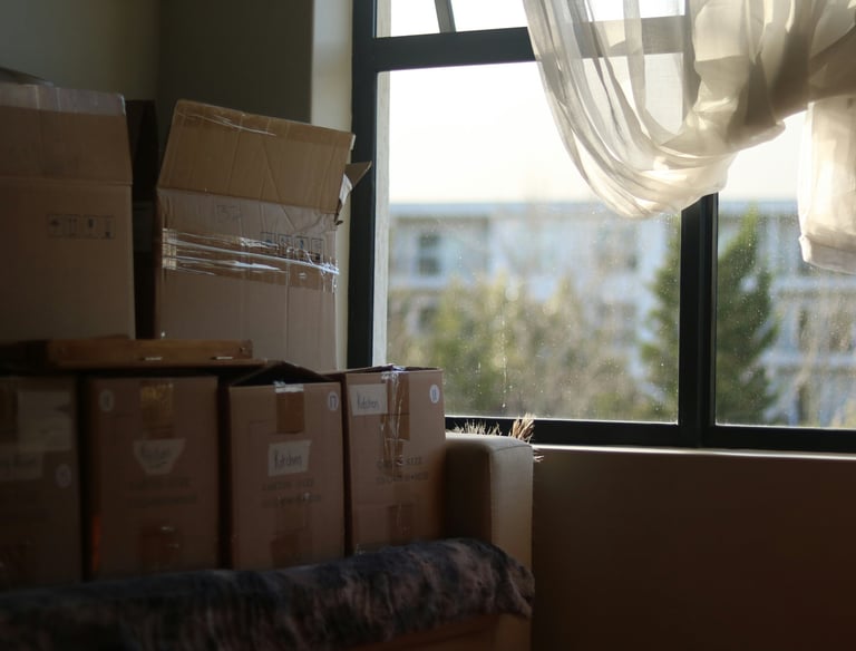 Stacked cardboard moving boxes in a bright room next to a window with white curtains.