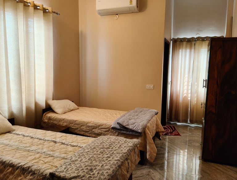 A bright PG room with two twin beds, tan curtains, and a wooden wardrobe on tiled floors.