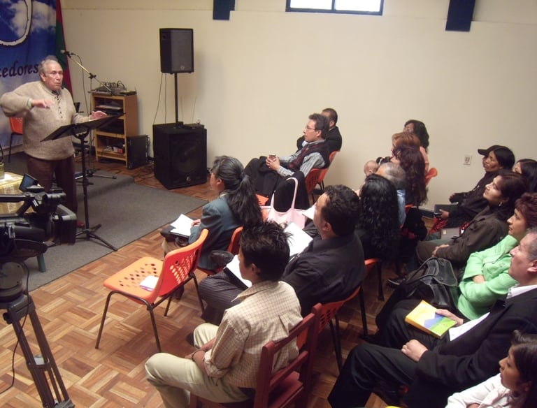 An elderly man speaking at a lectern during a community meeting or seminar with an attentive seated audience.