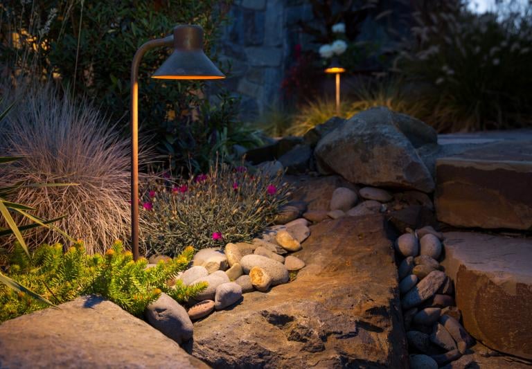 Low voltage landscape path lights illuminating a garden rockery and stone steps at dusk.