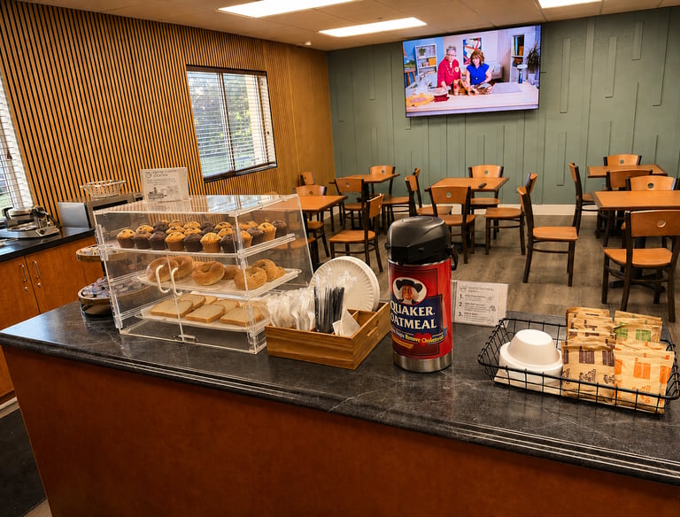 Hotel breakfast bar featuring fresh muffins, bagels, and a Quaker Oatmeal station in a dining area.