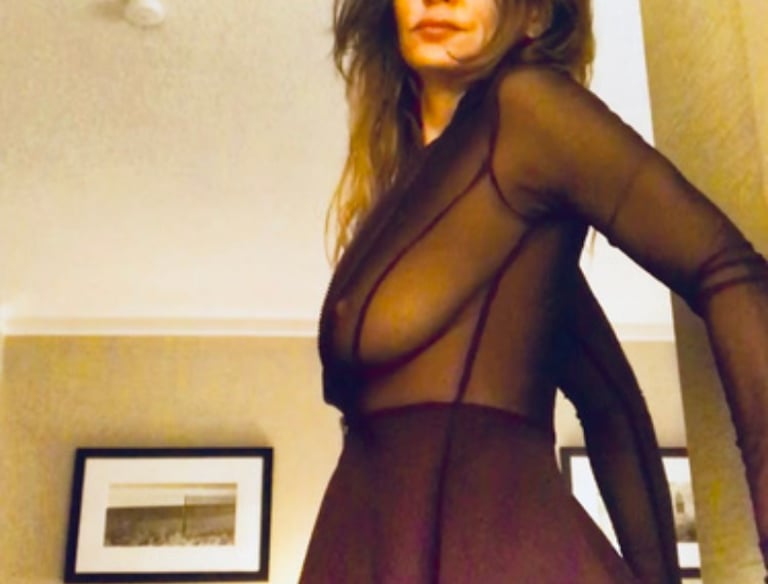 A woman posing in a dark sheer long-sleeve top and matching skirt in a room.
