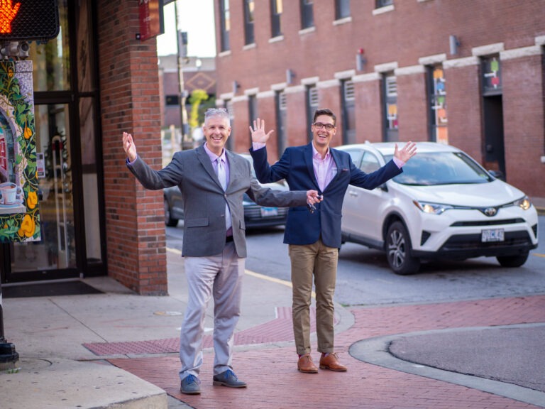 Lawrence and another man standing at a crosswalk with arms up smiling at the camera