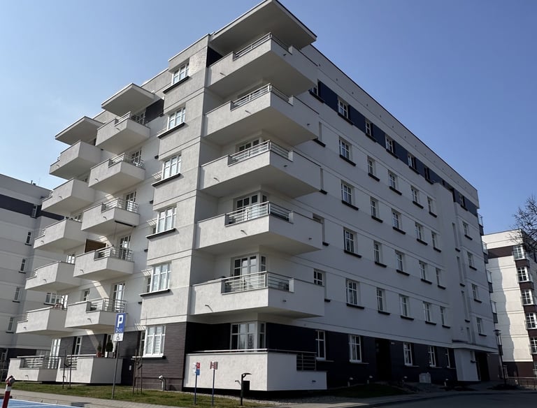 Nowoczesny budynek mieszkalny z jasną elewacją i szarą podstawą, z parkingiem z przodu.