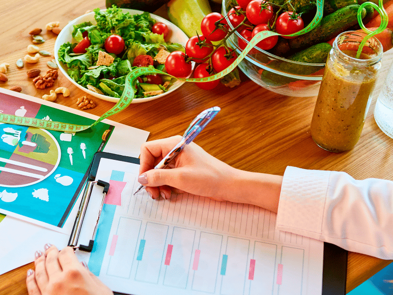 Mãos de nutricionista escrevendo em um prontuário sobre uma mesa com alimentos saudáveis