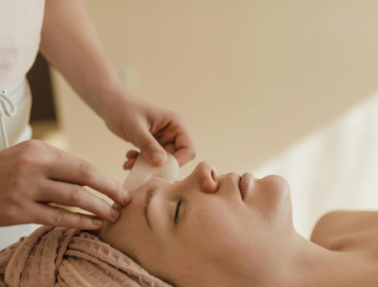 Mujer recibiendo masaje facial con piedra Gua Sha, relajada y con toalla en la cabeza en un ambiente
