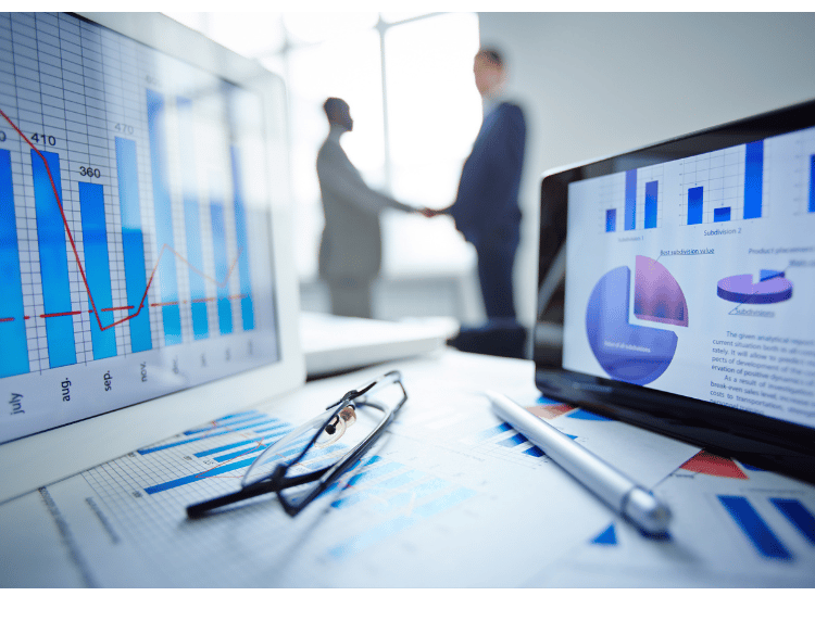 Financial data charts on a digital tablet and monitor with business people shaking hands in the background.