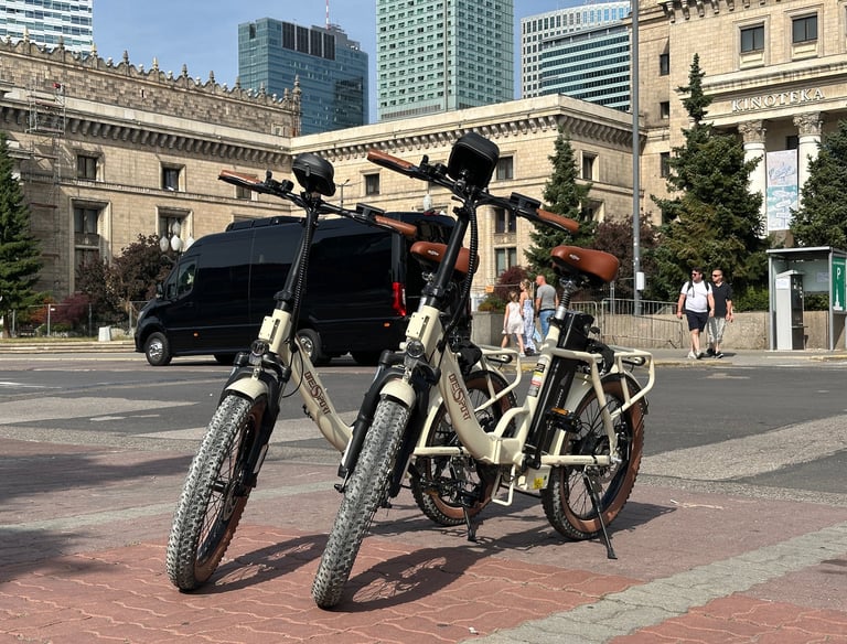 Two cream electric folding bikes with fat tires parked on a city street in Warsaw near modern skyscrapers.