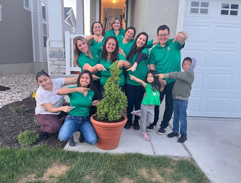 Smiling group in green shirts pointing at a potted boxwood shrub in front of a modern home.